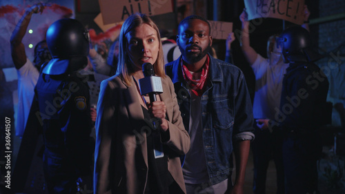 Beautiful serious woman journalist making a live speech on TV explaining the situation with night street demonstration rally. No racism. Black lives matter. Shocking news.