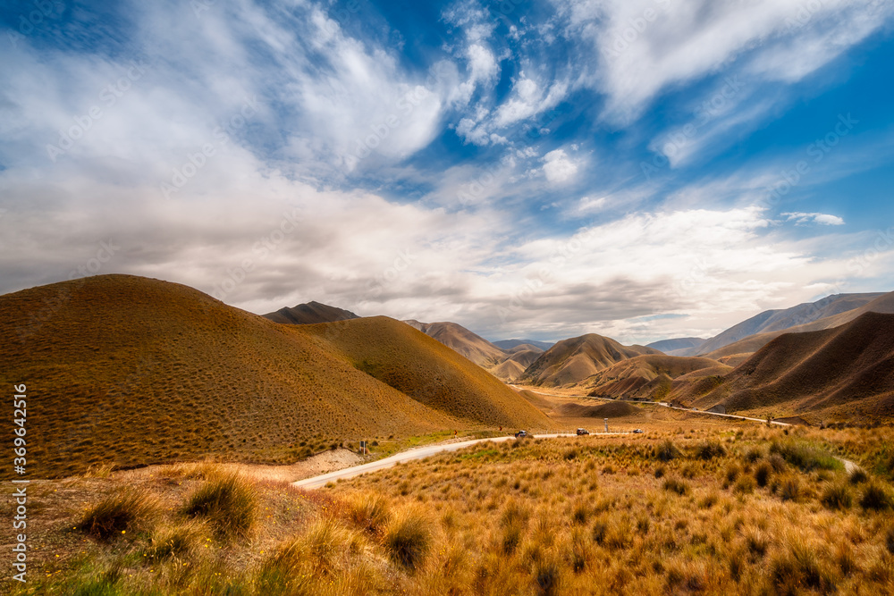Golden light at a New Zealand Desert Mountain Pass: A two-lane highway curves through arid terrain toward Lindis Pass in Otago Region, on a beautiful summer day in New Zealand, South Island.