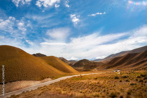 Golden light at a New Zealand Desert Mountain Pass: A two-lane highway curves through arid terrain toward Lindis Pass in Otago Region, on a beautiful summer day in New Zealand, South Island.