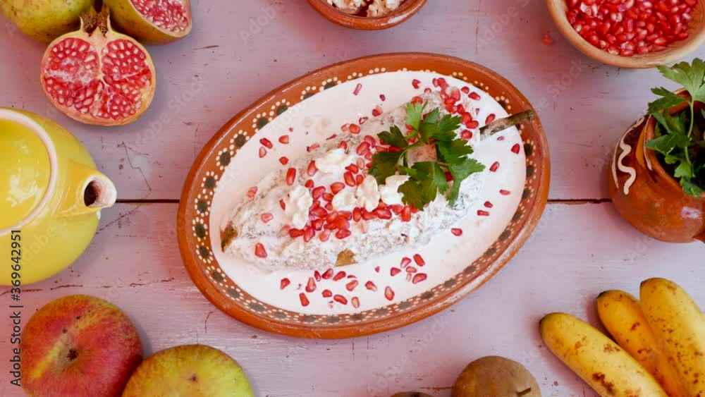 High angle view of chile en Nogada, typical Mexican dish in traditional clay plate with ingredients, pomegranate, bananas, apples, pears and parsley, panning motion on food