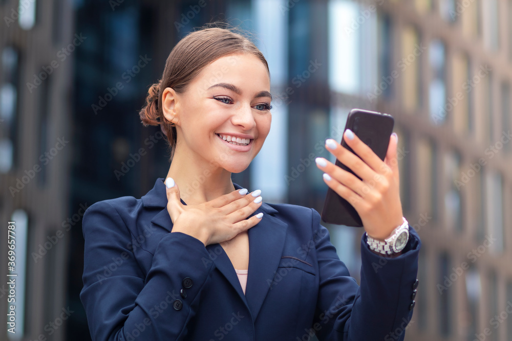 Happy excited beautiful businesswoman looking at screen of her mobile cell phone, smiling. Young woman having good news on smartphone. Girl in suit is reading nice message on telephone outdoors Happy excited beautiful businesswoman looking at screen of her mobile cell phone, smiling. Young woman having good news on smartphone. Girl in suit is reading nice message on telephone outdoors