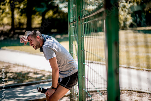 Young Caucasian man resting after outdoor run, leaning on fence.
