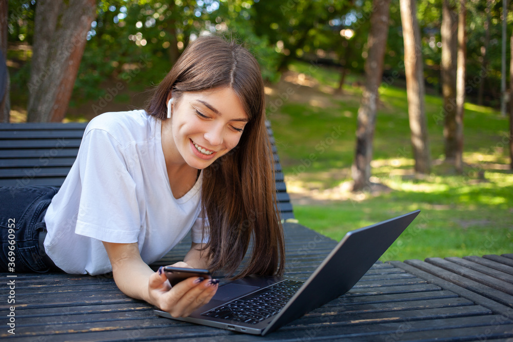 Remote working, education and technology concept. Young smiling beautiful student girl in the city park, wearing white t-shirt, in wireless earphones, using smartphone with laptop preparing for exams. Remote working, education and technology concept. Young smiling beautiful student girl in the city park, wearing white t-shirt, in wireless earphones, using smartphone with laptop preparing for exams.