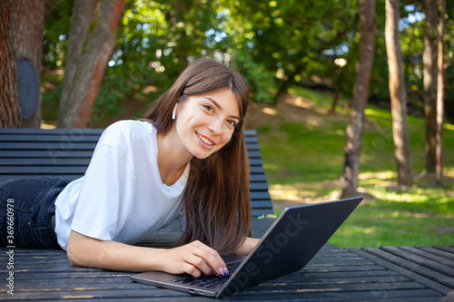 Remote working, education concept. Young smiling pretty student woman, lying on the bench in park, wearing wireless earphones, taking notes and making schedule on laptop preparing for exams. 