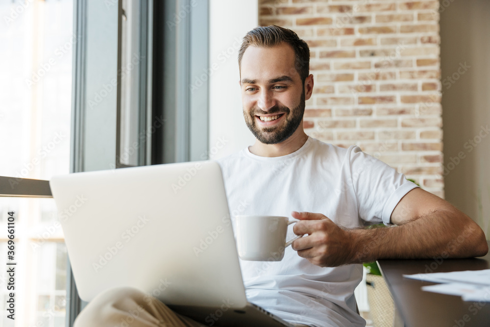 Image of man drinking coffee and using laptop while sitting at table Image of man drinking coffee and using laptop while sitting at table
