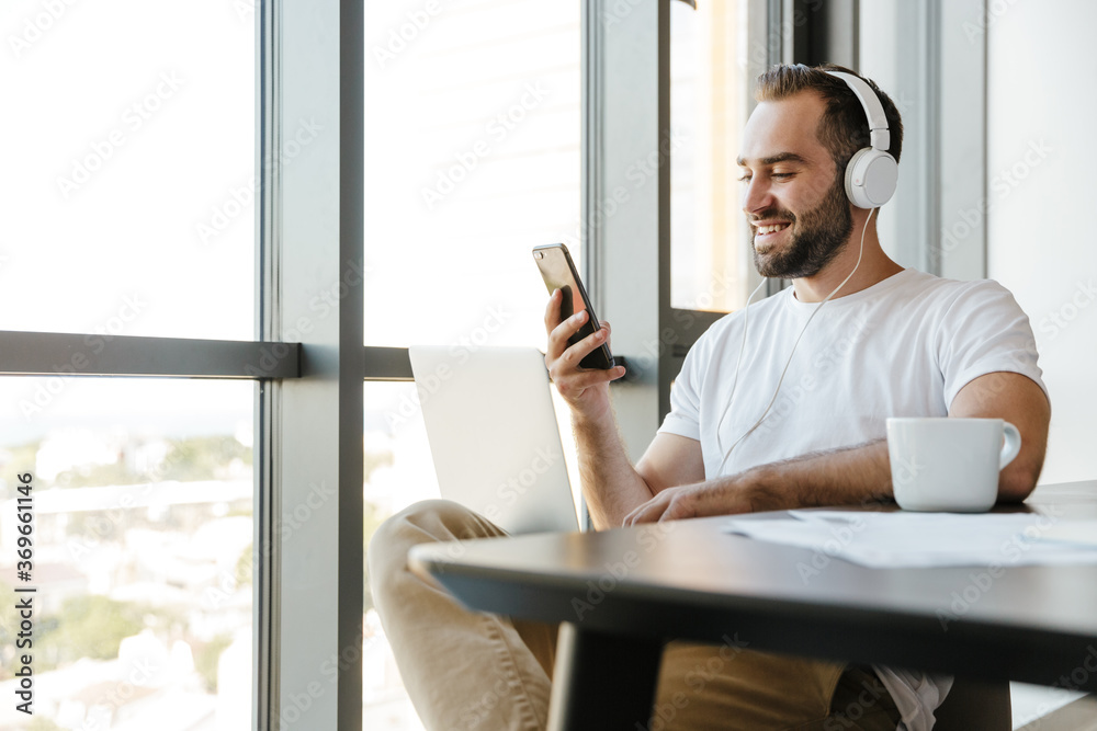 Image of smiling man using laptop and cellphone while sitting at table Image of smiling man using laptop and cellphone while sitting at table