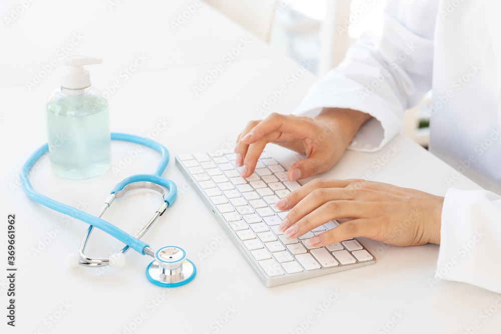 Closeup of female hands typing on computer keyboard, gel hand sanitizer and stethoscope on office desk. Online medical consultation and virus protection concept. Closeup of female hands typing on computer keyboard, gel hand sanitizer and stethoscope on office desk. Online medical consultation and virus protection concept.