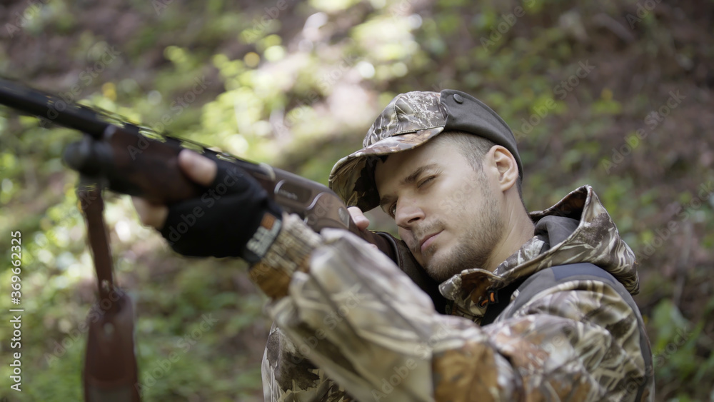 Man takes aim while standing from hunting rifle. Man in comfortable camouflage clothes hunter outdoor in forest hunting alone. Hunter in camouflage aims gun at object in forest.