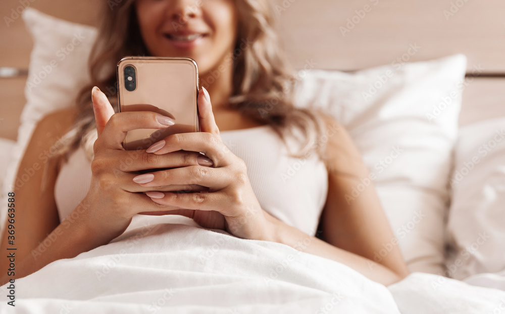 Young woman lying in bed holding a mobile phone in her hands, close up Young woman lying in bed holding a mobile phone in her hands, close up