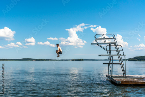 Sticker Side view of a teenage male jump diving from a diving tower with blue sky and horizon in the background