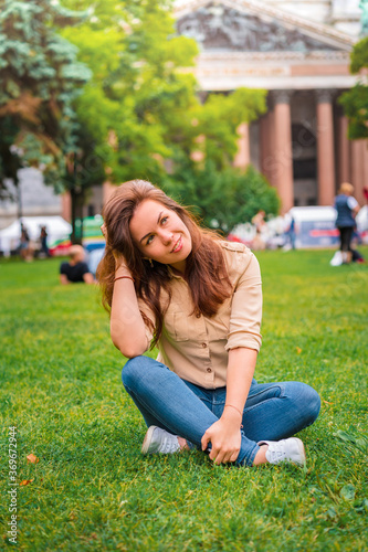 A cute girl with long brunette hair relaxes on the grass in the center of St. Petersburg in front of St. Isaac's Cathedral © KseniaJoyg