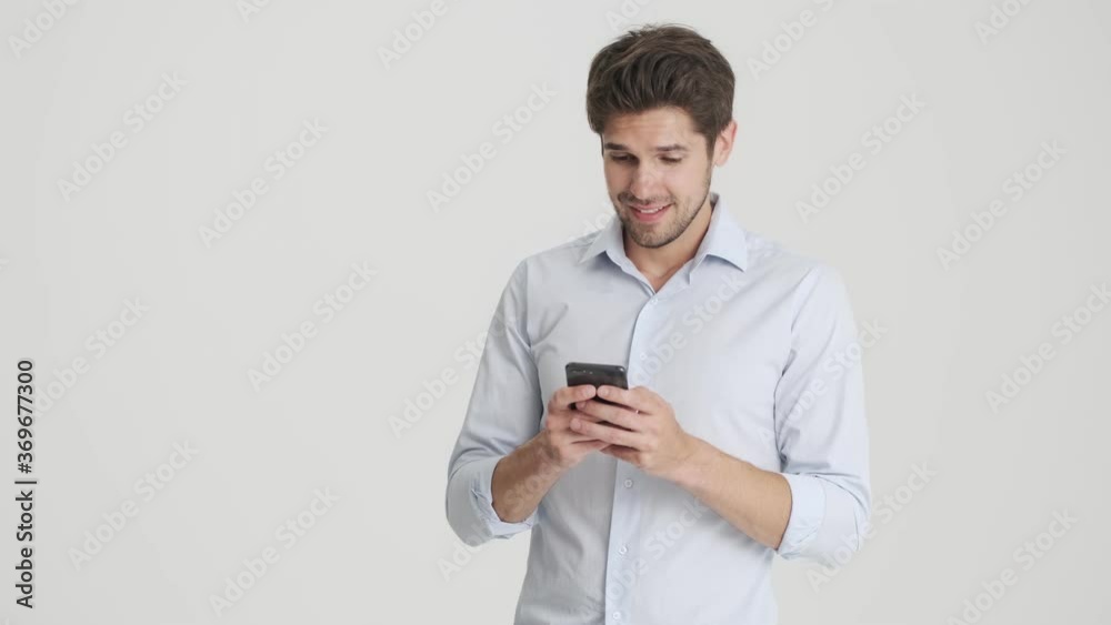 A happy man is using his smartphone while rejoicing and doing winner gesture standing isolated over white background A happy man is using his smartphone while rejoicing and doing winner gesture standing isolated over white background