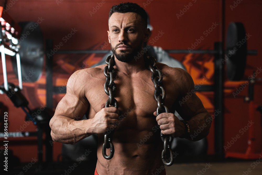 Portrait of a beautiful male bodybuilder with a bare torso and a thick chain hanging on his shoulders against the background of a heavy barbell and a red wall of the gym.