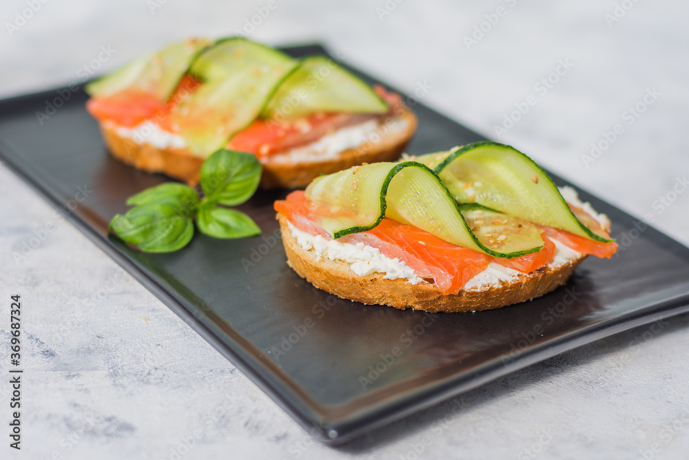 Open sandwiches with trout and cream cheese with cucumber and herbs on a dark plate - a tray on a gray table on a bright sunny day Open sandwiches with trout and cream cheese with cucumber and herbs on a dark plate - a tray on a gray table on a bright sunny day