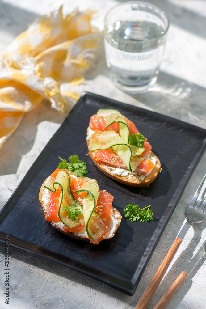 Open sandwiches with trout and cream cheese with cucumber and herbs on a dark plate - a tray on a gray table on a bright sunny day Open sandwiches with trout and cream cheese with cucumber and herbs on a dark plate - a tray on a gray table on a bright sunny day