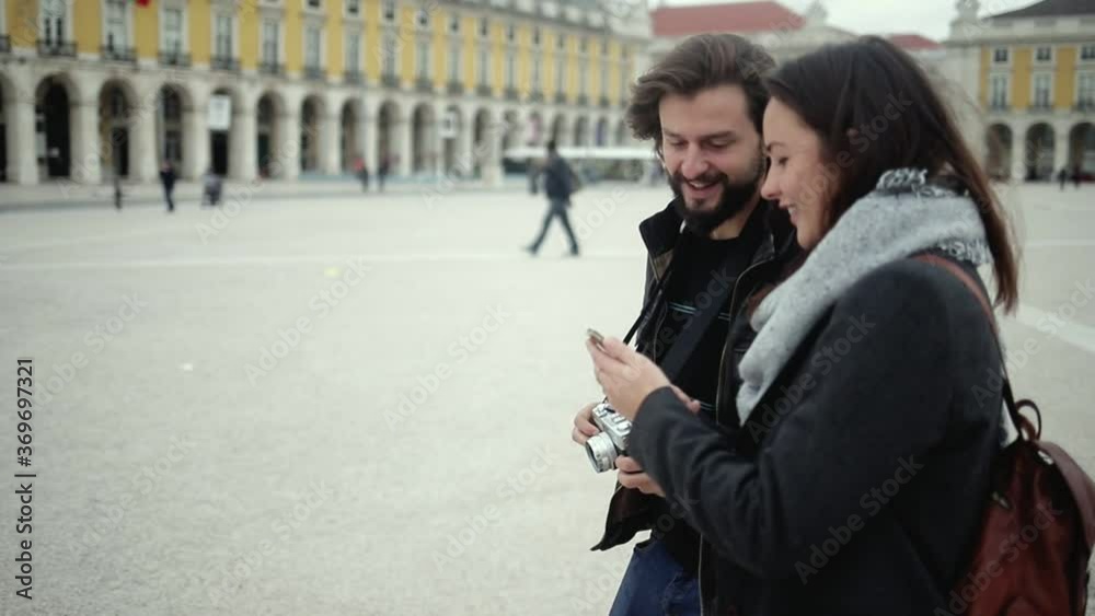 Young couple of tourists with cellphone and camera pointing hand at landmarks, taking pictures while walking on European city square. Medium shot, front view. Tourism concept Young couple of tourists with cellphone and camera pointing hand at landmarks, taking pictures while walking on European city square. Medium shot, front view. Tourism concept