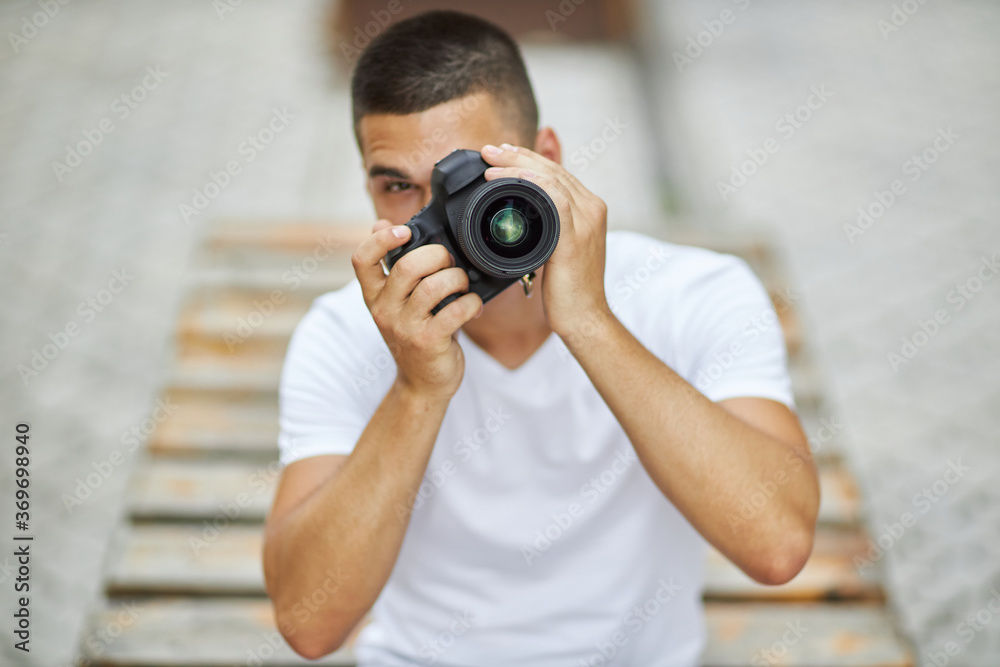 A young man is sitting outdoors with a camera and wants to take a photo. A young man is sitting outdoors with a camera and wants to take a photo.