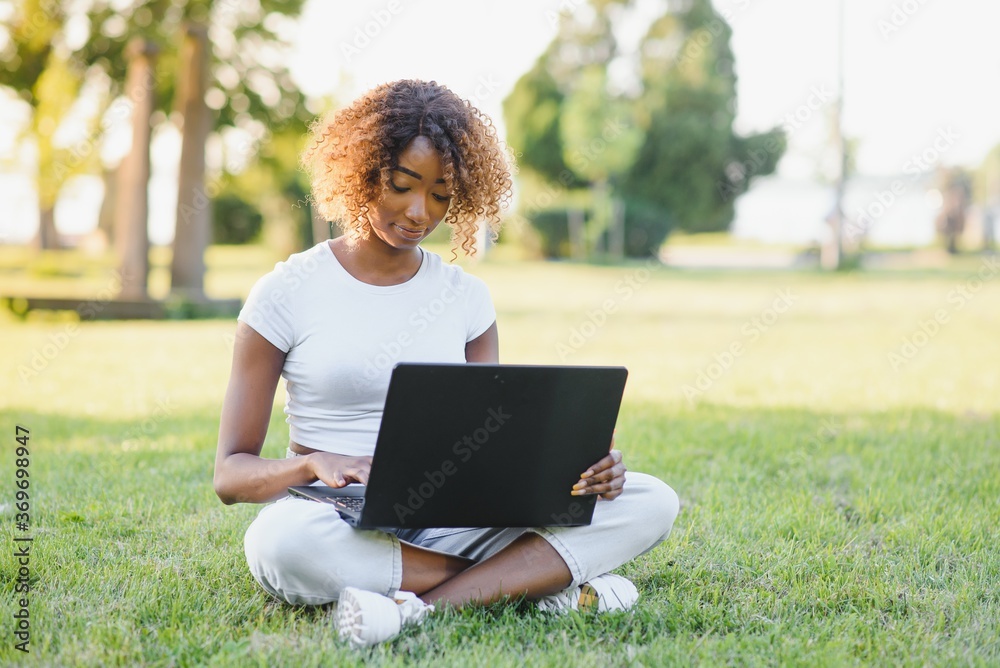 Young attractive dark-skinned college student wearing tank top and jeans sitting on the lawn at campus on sunny day, working on her thesis using laptop computer, looking busy and concentrated Young attractive dark-skinned college student wearing tank top and jeans sitting on the lawn at campus on sunny day, working on her thesis using laptop computer, looking busy and concentrated