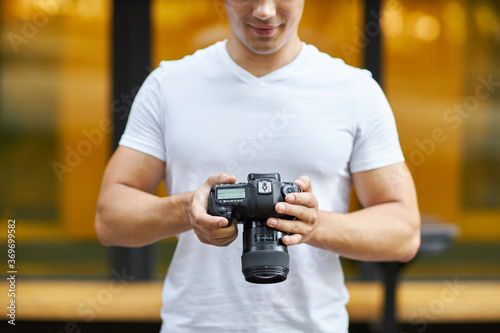 A young man stands outdoors with a camera and adjusts the camera