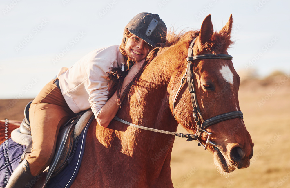 Fototapeta premium Young woman in protective hat with her horse in agriculture field at sunny daytime