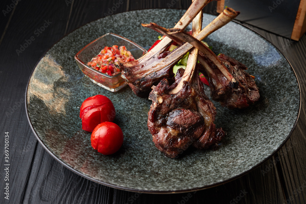 Rack of lamb on a dark plate with tomatoes and sauce, on a black background