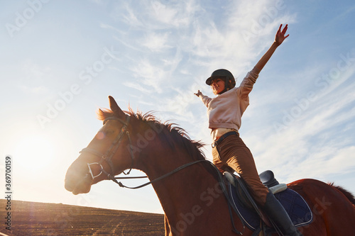 Fotografia Young woman in protective hat with her horse in agriculture field at sunny dayti