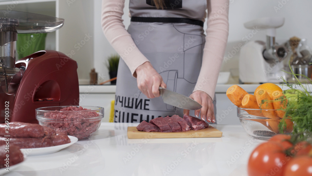 Woman cuts fresh meat standing at table in kitchen in bright house. Female cutting red beef filet on wooden board, holding knife in hand. Written on apron: best recipes