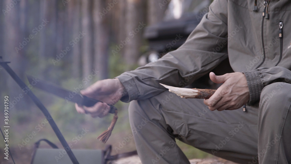 Male tourist is sitting on log and planing wooden peg with knife in summer forest. Cooking dinner on wild hike. Cauldron of hot tea or food is heated over fire. Outdoor travel.Boil of hot meal in camp