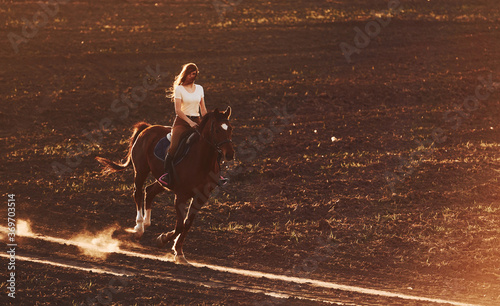 Young woman in protective hat riding her horse in agriculture field at sunny daytime