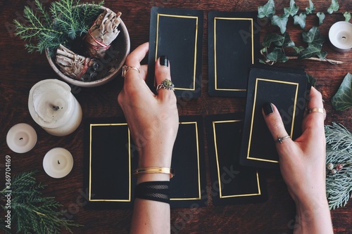 Wiccan witch doing a tarot card reading to predict the future. Female woman hands holding hand made tarot card deck. Black cards with gold rim. Nature evergreens, plants in dark wooden background