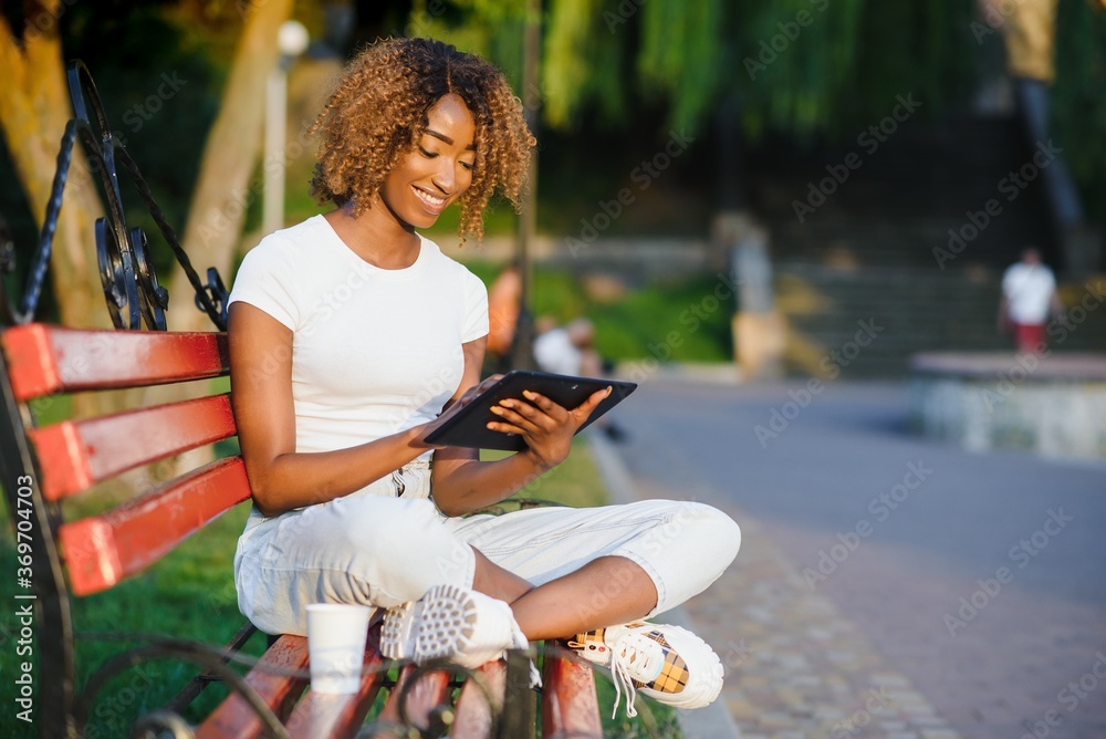 Fototapeta premium Young curly black smiling female with a digital pad on the bench in the park; charming cheerful Brazilian girl with Afro hair using tablet pc while sitting on the wooden bench on a summer day