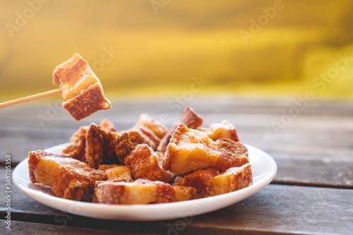A toothpick holding a piece of fried pork rinds called “Torreznos” over a small white round fried pork rinds plate on a dark wooden table. Photo with selective focus. 