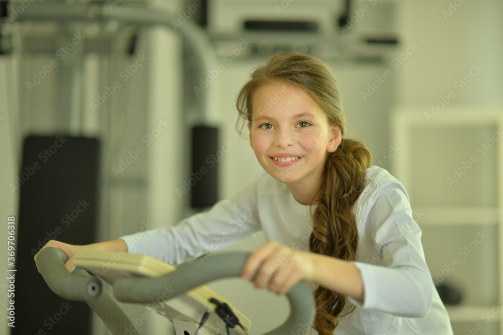 Fototapeta Little girl doing exercises on bike in gym