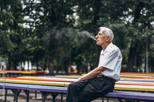 Senior adult men smoking cigarette outdoors in the city park when sitting on the bench.