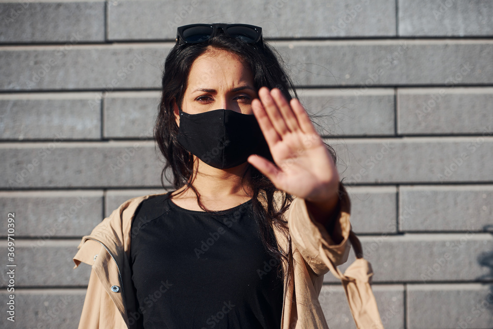 Naklejka premium Showing stop gesture by hand. In protective mask. Beautiful brunette with curly hair and in black clothes outdoors near wall