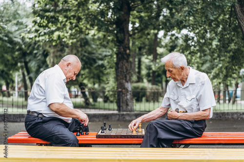 Two senior adult men playing chess on the bench outdoors in the park.