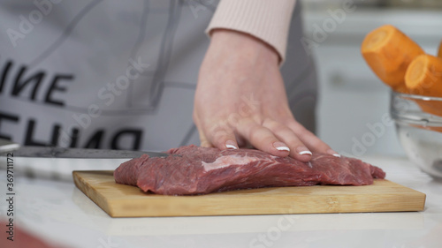 Fototapeta Naklejka Na Ścianę i Meble -  Woman cutting raw meat on small pieces with kitchen knife cutting board. Female cutting red beef filet on wooden board, holding knife in hand. Housewife is cooking at home interior. Concept: food