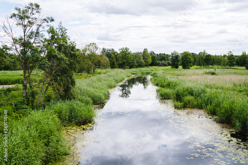 narrow river in a rural area, reflection of the sky and clouds in the water, grassy fields on both sides