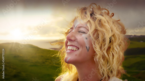 Beautiful, carefree smiling girl with blonde wavy hear and Celtic symbols painted on her face enjoying the sunset at the coast at dusk.
