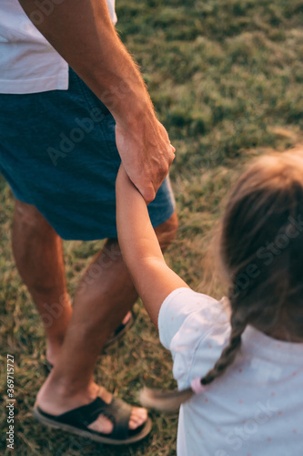 Father and daughter hold hands. The child's hand and the man's hand hold each other. Dad and daughter walk together holding hands on the grass in the summer at sunset