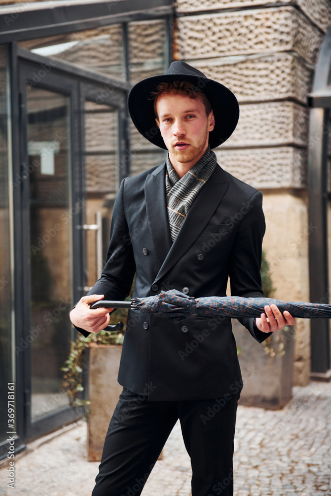 Obraz premium Elegant young man in black hat and clothes with umbrella in hands outdoors in the city