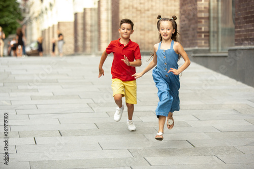 Two smiling kids, boy and girl running together in town, city in summer day. Concept of childhood, happiness, sincere emotions, carefree lifestyle. Little caucasian models in bright clothes.