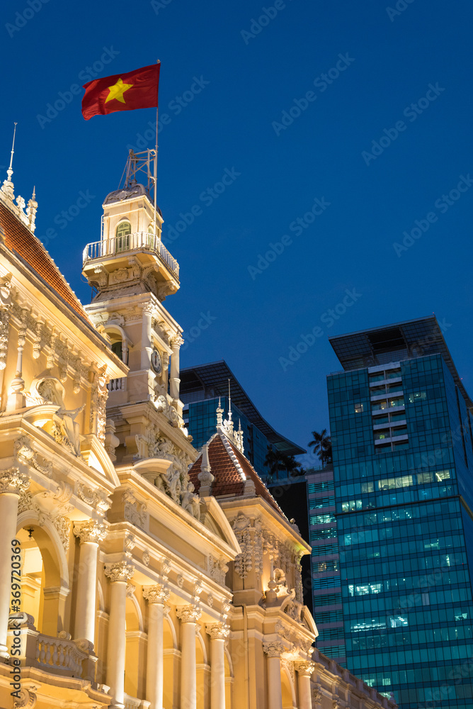 Obraz premium Saigon City Hall (People's Committee Head office) with waving Vietnamese flag at night. Ho Chi Minh City, Vietnam. Vertical photo