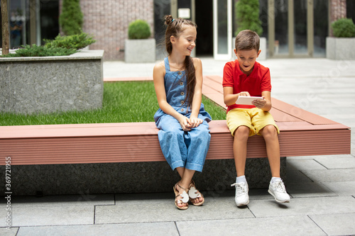Two smiling kids, boy and girl playing smartphone in town, city in summer day. Concept of childhood, happiness, sincere emotions, carefree lifestyle. Little caucasian models in bright clothes.