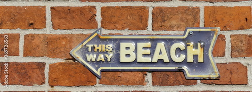 This way beach. Old rusty metal sign on a red brick wall. Blue with white lettering. Path to the beach, signpost and tin sign with an arrow pointing to the left.