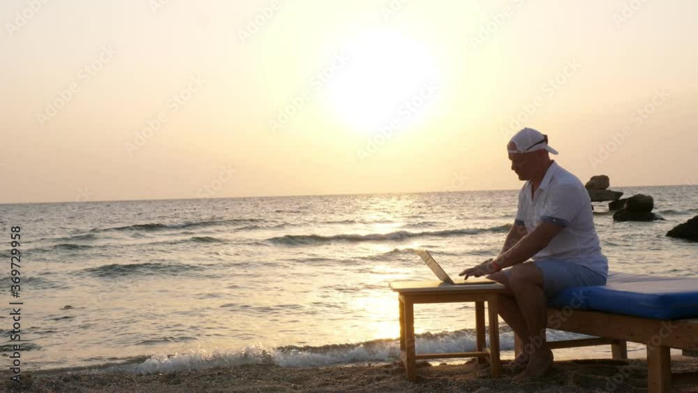 man in blazer hat, typing using laptop, sitting on the beach by the sea, at sunset or sunrise. freelancer, remote work. summer vacation at sea. internet erases distances man in blazer hat, typing using laptop, sitting on the beach by the sea, at sunset or sunrise. freelancer, remote work. summer vacation at sea. internet erases distances