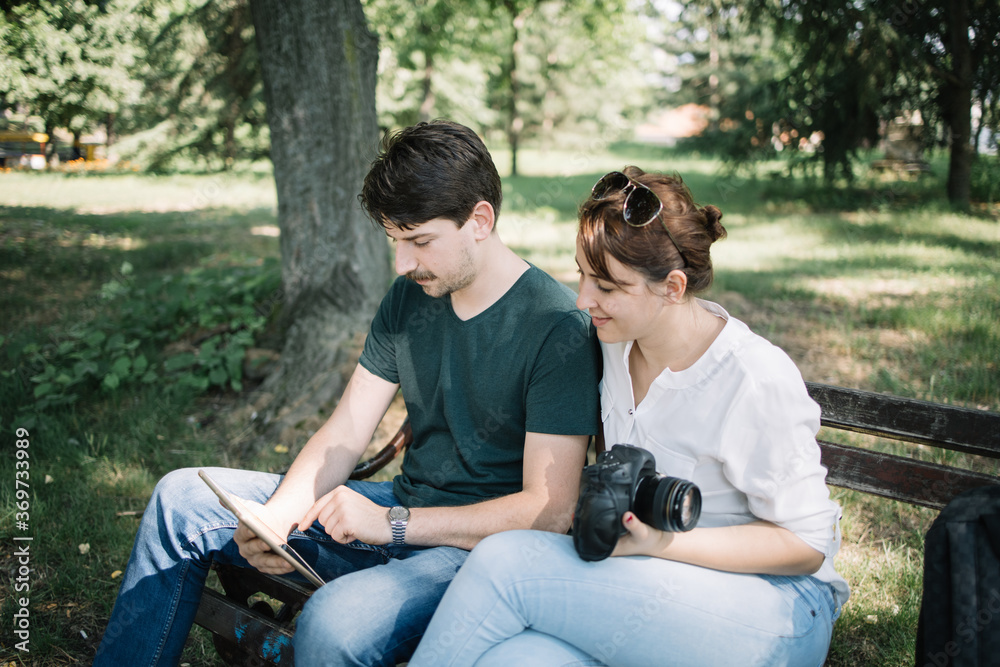 Photographer and model checking photo session photos on tablet. Happy young couple sitting on bench and looking at tablet in park. Photographer and model checking photo session photos on tablet. Happy young couple sitting on bench and looking at tablet in park.