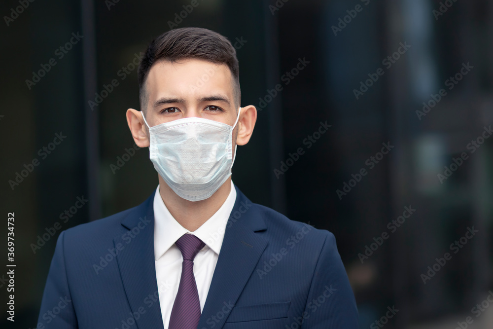 Portrait of handsome serious businessman in medical mask on his face, young man, guy in suit, jacket, shirt, tie looking at camera outdoors. Coronavirus, covid-19, pandemic, virus, business concept Portrait of handsome serious businessman in medical mask on his face, young man, guy in suit, jacket, shirt, tie looking at camera outdoors. Coronavirus, covid-19, pandemic, virus, business concept
