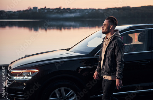 Handsome unshaved man in fashionable clothes walks near his black car at evening time