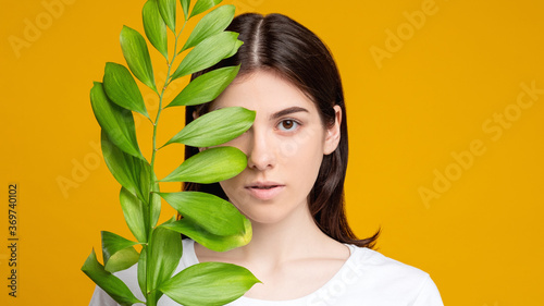 Natural beauty. Skin care. Portrait of woman covering fresh clean face with exotic green leaf isolated on orange copy space. Aesthetic cosmetology. Organic mask.